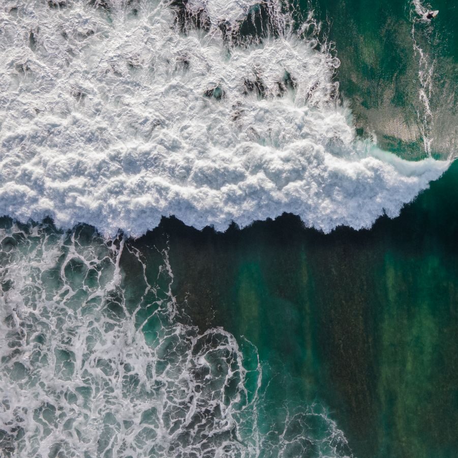 Aerial view of ocean wave breaking over deep turquoise water.